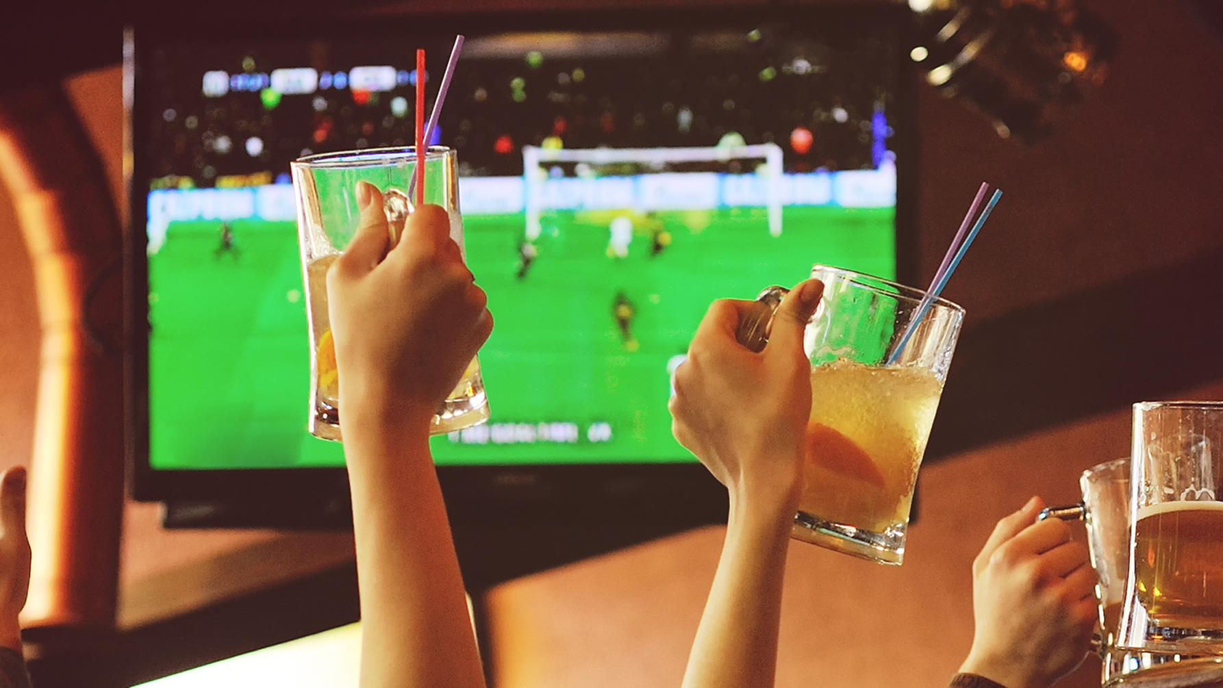 Sports fans hands holding beer glasses in front of a TV screen celebrate