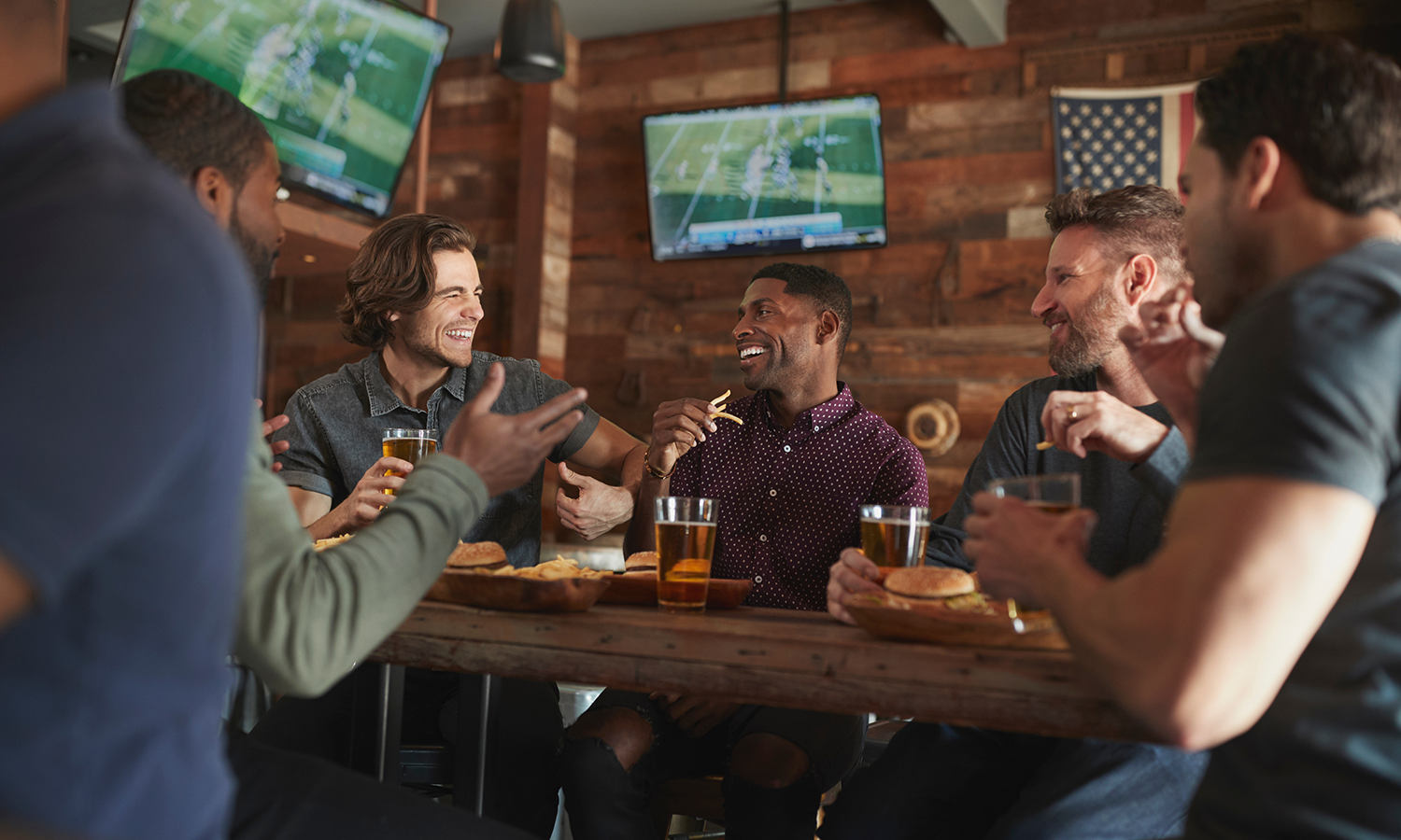 Group of young male sports fans watching professional football on TV while chatting and enjoying beer & apps