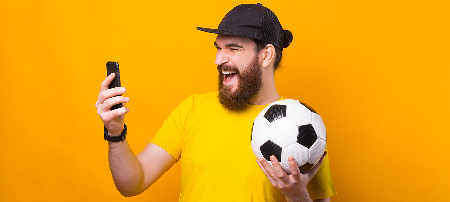 Male sports fan in a yellow shirt and black hat holding a soccer ball and smiling while reading an SMS marketing message from a local sports bar